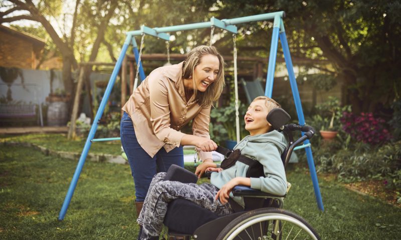Cheerful woman removing strap from happy special young son in wheelchair in garden Woman wearing casual attire, leaning forward smiling cheerfully, calmly removing strap from happy young special son sitting in wheelchair, enjoying morning air and sun in garden